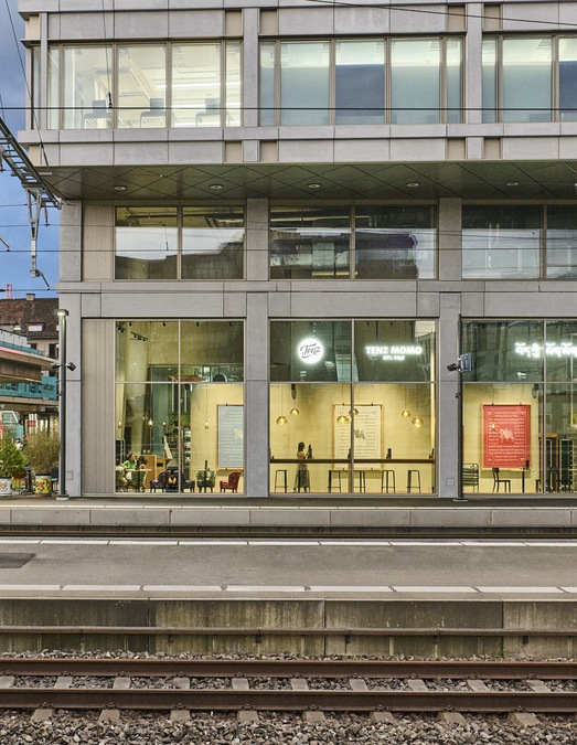 Nächtliche Aussenansicht des Bahnhofsgebäudes Zürich Oerlikon mit beleuchtetem Schriftzug Tenz Momo hinter grossen Fensterfronten, im Vordergrund Gleise und Bahnsteig